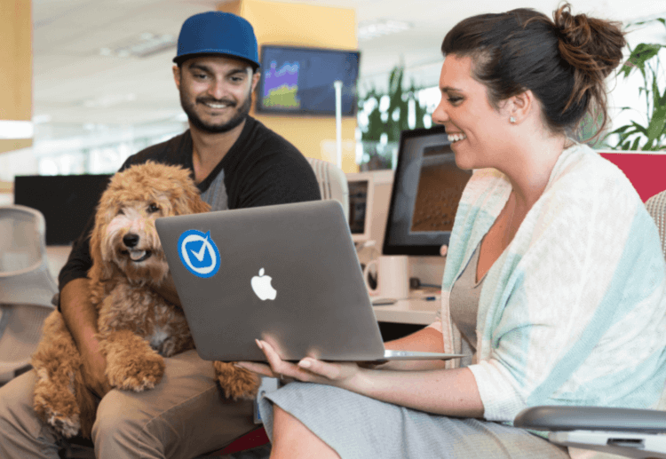 Two smiling colleagues collaborating on a laptop in the pet-friendly Clio office, with one holding a fluffy dog on their lap.