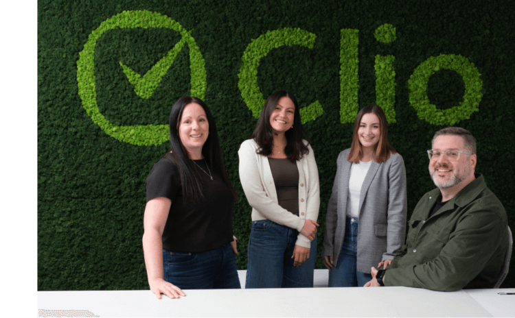 Group of four smiling employees in front of a green moss Clio logo wall.