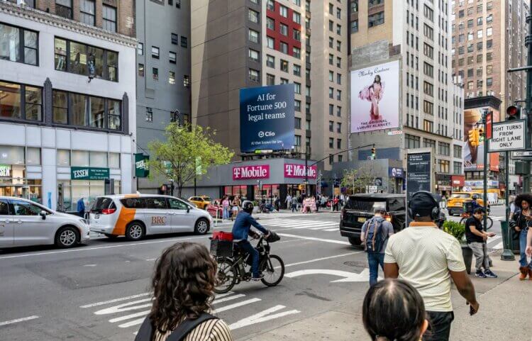 A busy NYC street corner with a digital billboard reading "AI for Fortune 500 legal teams" by Clio above a T-Mobile store.
